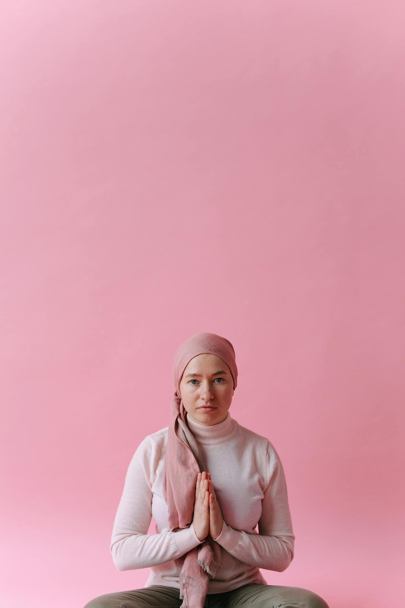 A woman in a headscarf sits gracefully, embodying hope and strength against a soft pink background.